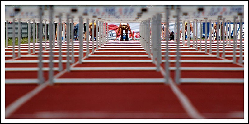 Athletes at the start of a hurdles race. The image looks down the track, through the gaps under a series of hurdles to the athletes.