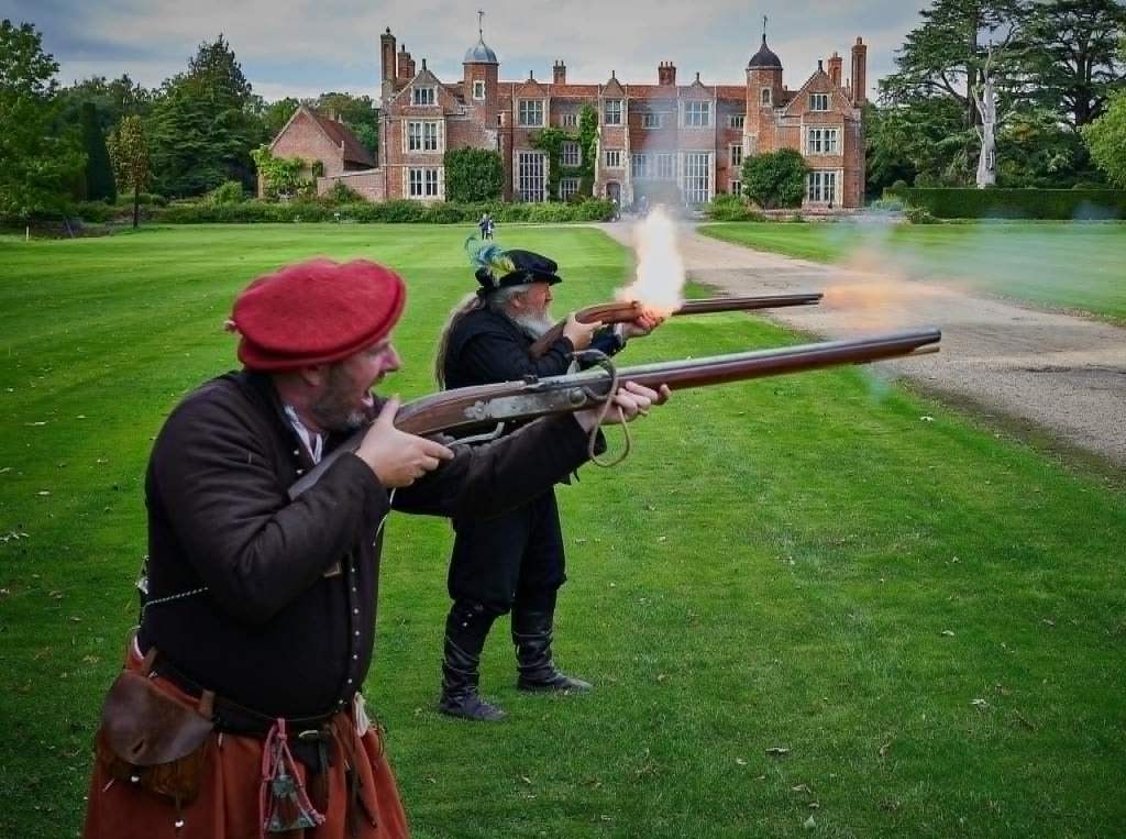 Two men, dressed as Tudors, shoot replica calivers (long guns) on the front lawn of Kentwell Hall, with the hall in the background